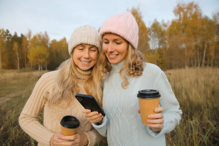 Happy modern young two women friends with smartphone together in autumn park, smiling girlfriends using phone with cup of coffee drinks wearing winter hat, sweater outdoors on a warm dayの写真素材