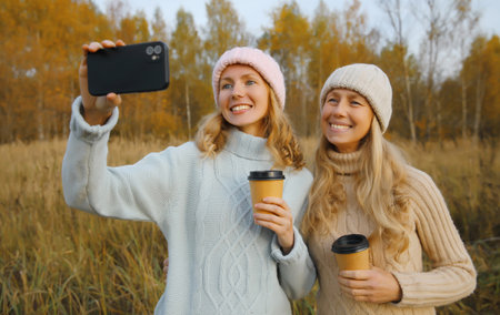 Happy two women friends taking selfie with phone in autumn park, joyful girlfriends smiling having fun together with cup of coffee drink outdoors wearing winter hat, sweater in warm fall seasonの写真素材