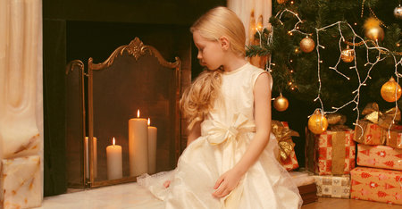 Happy little girl child in dress with festive gift box sitting near Christmas tree and fireplace with holiday lights at homeの写真素材