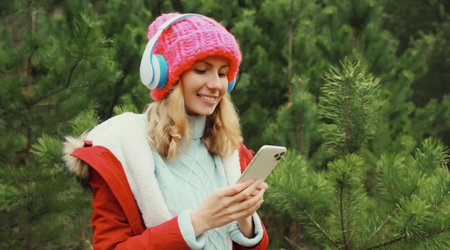 Portrait of happy modern young woman with phone enjoying listening to music in headphones in hat,...