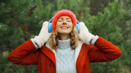 Portrait of happy modern young woman enjoying listening to music with headphones in hat, jacket in winter forest against Christmas tree background with snowの写真素材
