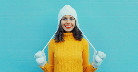 Winter christmas portrait of happy young woman smiling in yellow sweater, white hat standing on blue backgroundの写真素材