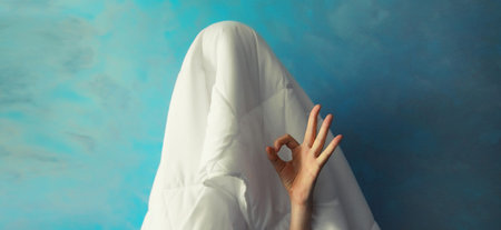 Cheerful lazy woman waking up after sleeping covered head with white blanket showing ok gesture her fingers on blue backgroundの写真素材