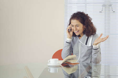 Smiling happy woman with coffee and a book, sitting at a table and talking on the phone.の写真素材