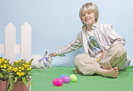 Blonde seated boy reaches out to pet a white rabbit wearing a bow