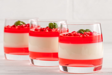 Creamy panna cotta with red jelly in beautiful glasses, fresh ripe pomegranate seeds on white wooden background. Delicious Italian dessert. Close up photography. Selective focus. Horizontal banner.の写真素材