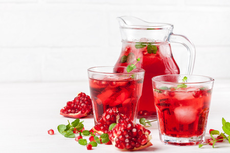 Pomegranate cocktail with ice and mint in beautiful glasses and jug, fresh ripe pomegranate on white wooden background. Sweet red juice. Close up photography. Selective focus. Horizontal banner.の写真素材