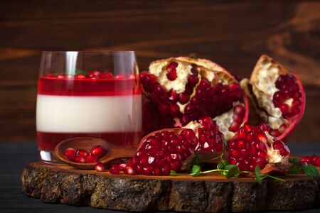 Creamy panna cotta with red jelly in beautiful glasses, fresh ripe pomegranate on wooden background.の写真素材