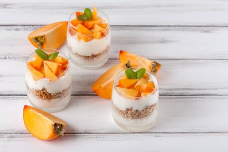 Persimmon creamy trifle in beautiful glasses, fresh ripe fruit slices on white wooden background. Healthy vegetarian food. Delicious dessert. Close up photography. Selective focus. Horizontal banner.の写真素材