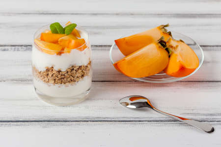 Persimmon creamy trifle in beautiful glasses, fresh ripe fruit slices on white wooden background. Healthy vegetarian food. Delicious dessert. Close up photography. Selective focus. Horizontal banner.の写真素材