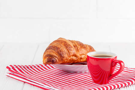 Freshly baked croissant on napkin, cup of coffee in red cup on white wooden background. French breakfast. Fresh pastries for breakfast. Delicious dessert. Closeup photography. Horizontal banner.の写真素材