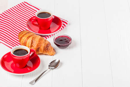 Freshly baked croissant on napkin, jam, cup of coffee in red cup on white wooden background. French breakfast. Fresh pastries for breakfast. Delicious dessert. Closeup photography. Horizontal banner.の写真素材