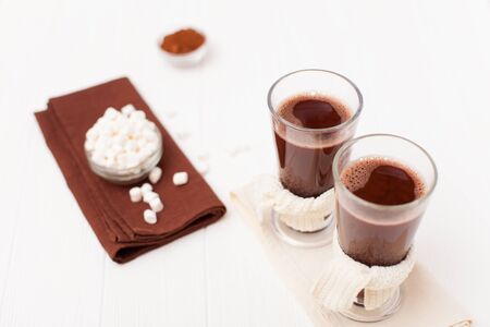 Warm cocoa in glasses with marshmallow, crocahet decorations, brown and beige napkins on white wooden background. Hot chocolate. Sweet drink. Close up photography. Hoizontal banner.の写真素材