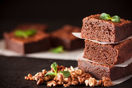 Chocolate brownie square pieces in stack on white plate with walnuts, decorated with mint leaves and cocoa on black background. Delicious dessert. Dark mood. Close up photography. Selective focus.の写真素材