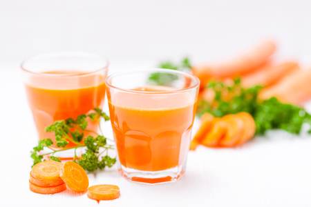 Carrot juice in beautiful glasses, cut orange vegetables and green parsley on white wooden background. Fresh orange drink. Close up photography. Selective focus. Horizontal banner.の写真素材