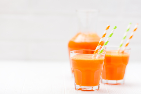 Carrot juice in beautiful glasses, cut orange vegetables and green parsley on white wooden background. Fresh orange drink. Close up photography. Selective focus. Horizontal banner.の写真素材
