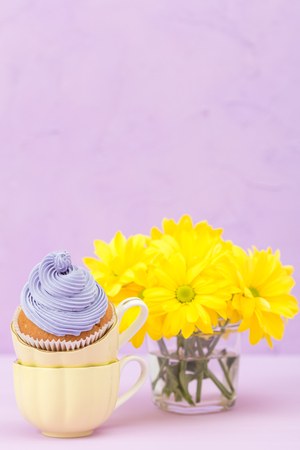 Cupcakes decorated with violet cream and bouquet of yellow chrysanthemums on violet pastel background. Text area. Ideal for greeting, mothers days and valentines card. Minimal still life concept.の写真素材