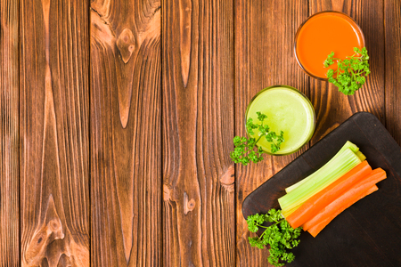 Carrot and celery juice in glasses with cut fresh vegetables and green parsley leaves on kitchen board with copy space on wooden background - top view of fresh raw healthy organic vegetarian food.の写真素材