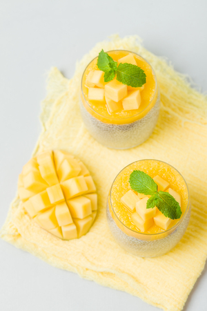 Chia seeds pudding with mango puree in beautiful glasses with green mint leaves and cut fresh ripe tropical fruit on light gray background - raw vegetarian sweet organic dessert.の写真素材