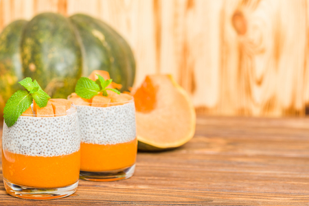 Chia pudding with pumpkin puree in beautiful glasses with green mint leaves and fresh ripe vegetable on brown wooden background - raw vegetarian sweet healthy organic dessert.の写真素材