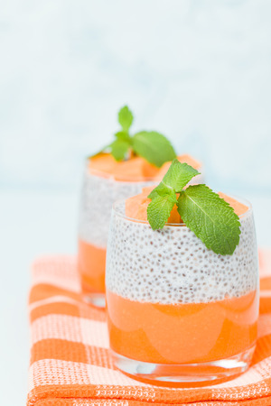 Chia seeds pudding with pumpkin puree in beautiful glasses with green mint leaves and cut fresh ripe orange vegetable on pastel blue background - raw vegetarian sweet organic dessert.の写真素材