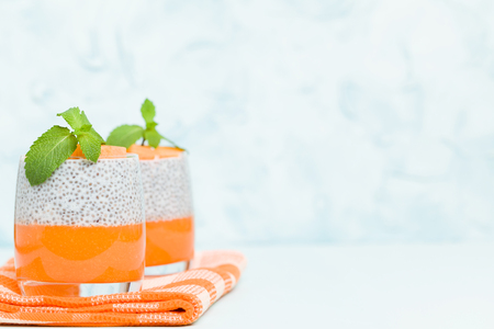 Chia seeds pudding with pumpkin puree in beautiful glasses with green mint leaves and cut fresh ripe orange vegetable on pastel blue background - raw vegetarian sweet organic dessert.の写真素材