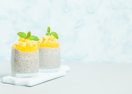 Chia seeds pudding with mango puree in beautiful glasses with green mint leaves and cut fresh ripe tropical fruit on pastel blue background - raw vegetarian sweet organic dessert.の写真素材
