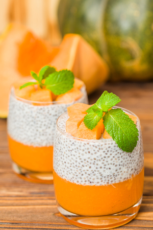 Chia pudding with pumpkin puree in beautiful glasses with green mint leaves and fresh ripe vegetable on brown wooden background - raw vegetarian sweet healthy organic dessert.の写真素材