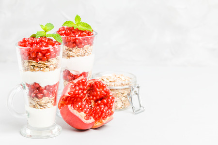 Pomegranate parfait - sweet organic layered dessert with granola flakes, yogurt and ripe fruit seeds in beautiful glasses on gray background with copy space. Natural vegetarian healthy food.の写真素材