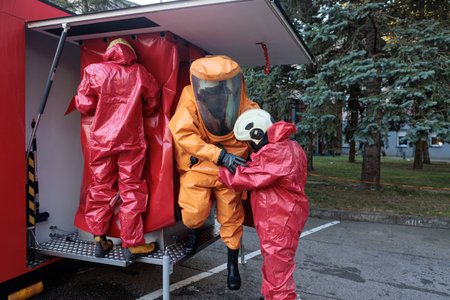 Emergency team members in red chemical suits helping a third responder in an orange hazmat suit enter a mobile decontamination unit or vehicle.の写真素材