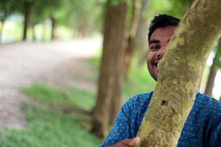 Bangladesh, Rangpur- August 16, 2021. A boy is standing behind a tree wearing blue Punjabi and the green natural background behind him is blurryのeditorial素材