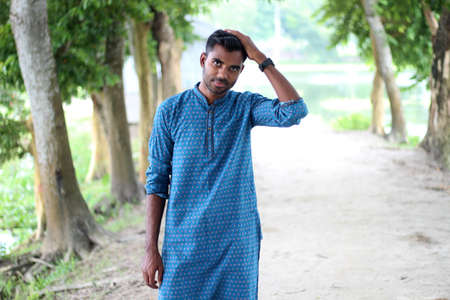Bangladesh, Rangpur- August 16, 2021. A young boy is standing in style wearing a blue Punjabi and show his hand watch and the green natural background behind him is blurryのeditorial素材