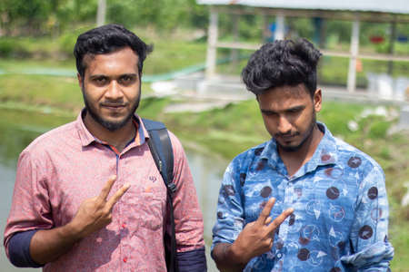 Bangladesh, Rangpur- August 21, 2021. Two young men are standing in the scorching sun and showing two fingers on one of their hands. his small beard and his behind blur the green nature background.のeditorial素材