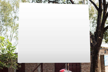 A big white signboard attaches two iron poles under a big tree and the background blur behind it.の写真素材