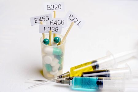 Unhealthy food: In a glass with multi-colored pills are plates with the code E-supplements. Nearby are syringes with nitrates, chemicals and additives. On the table is a flask with nitric acid - the main nitrate.の写真素材