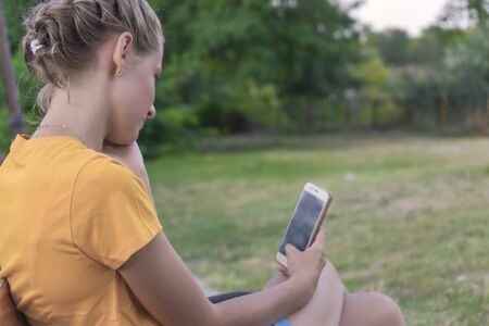 Girl with a phone in her hands, sitting on a bench in the park. Back view. Online shopping concept.の写真素材