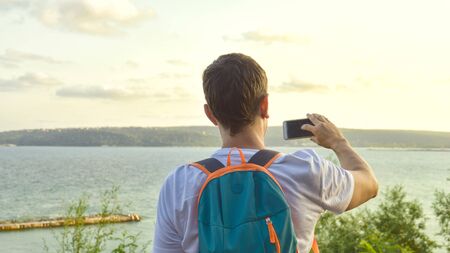 Journey through life concept. A male tourist stands on a viewing platform, admiring the sea view of the resort town, with a backpack and taking photos on a smartphoneの写真素材