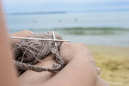 woman knits grey yarn openwork pattern with metal knittings on beach.の写真素材