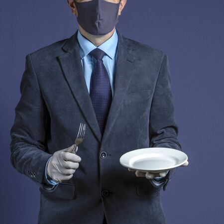 Photo businessman in a medical mask and medical gloves, holds in his hand an empty white plate and fork, isolated on background. Business crisis during a pandemic.の写真素材