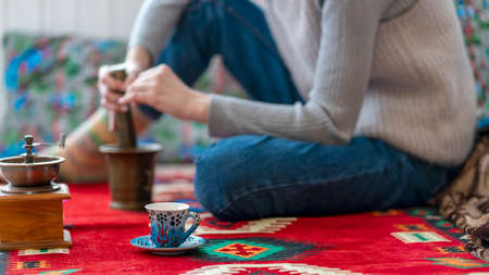 Young woman makes coffee at home. Sitting on an oriental rug, using a manual coffee grinder to make...
