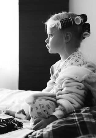 The girl is sitting on the bed. Little girl with curlers in the bedroom. Beauty and fashion concept.の写真素材