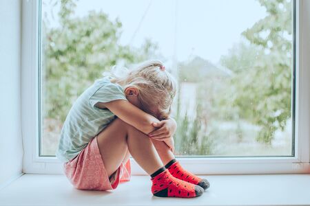 Little blonde girl sitting at the floor embracing her knees, near window at home, her head down, bored, troubled with parents fighting.の写真素材