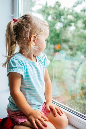 A gorgeous blonde little girl with ponytale staring out of the window on a wet, cold rainy summer day.の写真素材