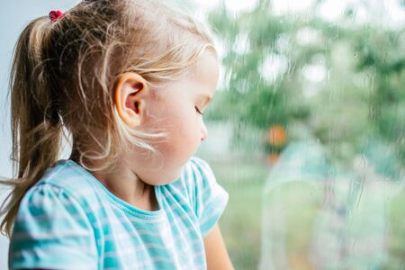 A gorgeous blonde little girl with ponytale staring out of the window on a wet, cold rainy summer day.の写真素材