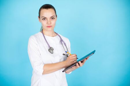 Young smiling female blonde doctor with phonendoscope on her neck and tablet on light blue...