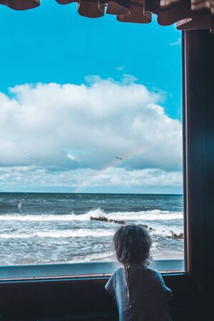 Lone little girl standing on window in front of the sea and looking to it. Sky is blue and waves on a seaの写真素材