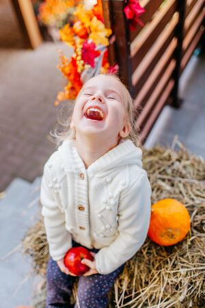 Adorable blonde baby toddler in white knittes jacket sitting on the haystack with pumpkins at porch, playing with apple and laughing. Halloween Thanksgiving card.の写真素材