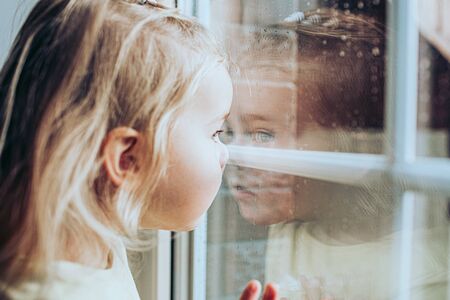 Little adorable blonde toddler girl looking through a window with rain drops on it. Close up...