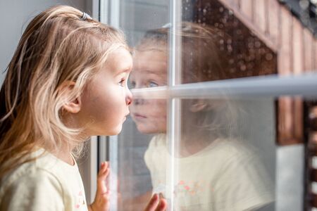 Little adorable blonde toddler girl looking through a window with rain drops on it. Close up portraitの写真素材