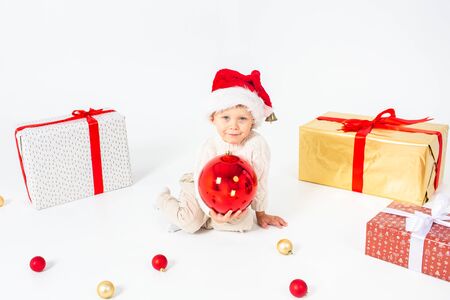 Little boy in Santas hat sitting between gifts and holding big red Christmas ball in hands. Isolated on white background.の写真素材